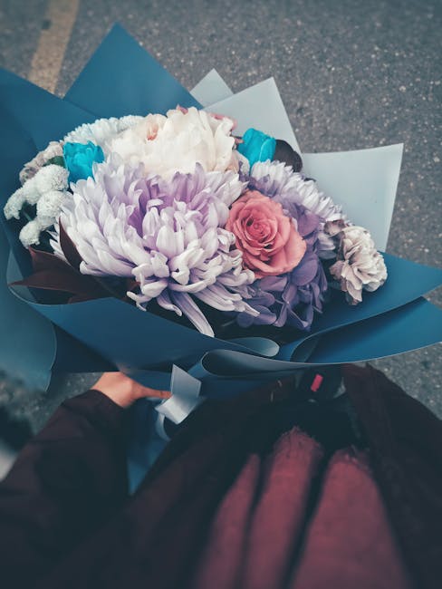 A floral bouquet featuring large, soft purple and white chrysanthemums, complemented by a single pink rose, with smaller white and purple flowers arranged throughout. The flowers are wrapped in dark b
