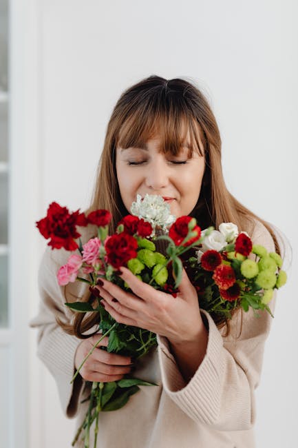 A woman with light brown hair and a beige sweater holds a vibrant bouquet of fresh flowers close to her face, with a gentle smile and her eyes closed. The bouquet includes red carnations, pink roses, 