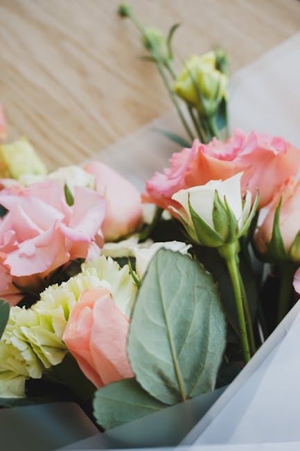 A close-up view of a floral arrangement featuring soft pink and cream lisianthus flowers with ruffled petals, accented by fresh green leaves, all wrapped in white paper with a grey ribbon. The flowers
