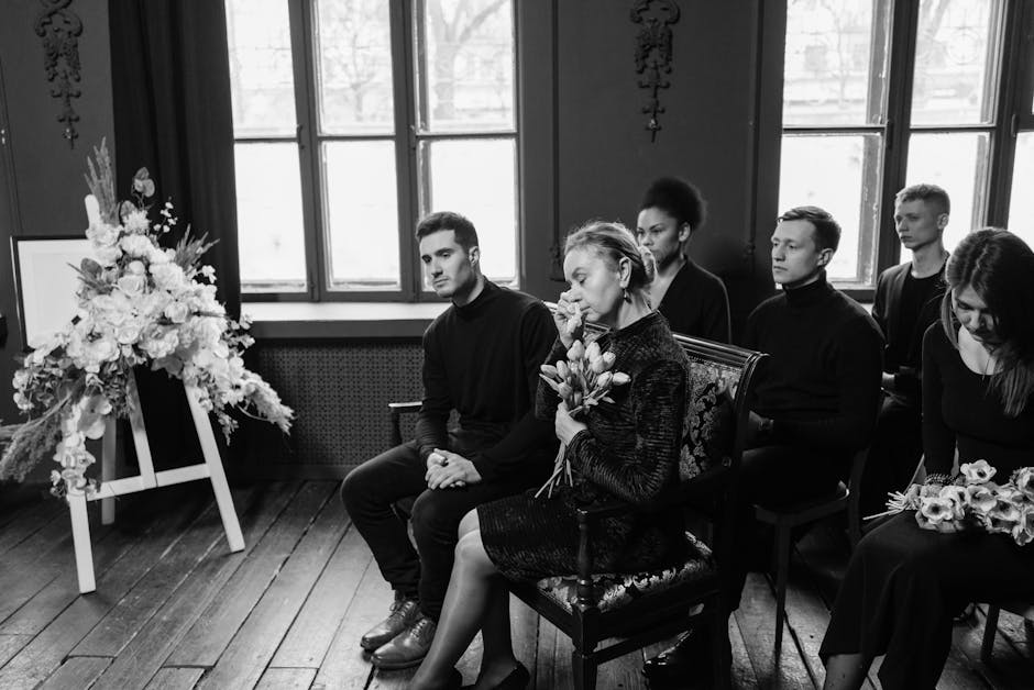 A black-and-white photograph capturing a solemn funeral scene inside a room with large, multi-paned windows allowing natural light. In the foreground, a woman is seated holding a floral bouquet consis