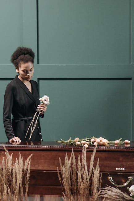 A young woman with dark curly hair, dressed in a black blazer, stands beside a wooden casket in a funeral setting. She holds a small white floral arrangement with a beige ribbon, and her gaze is direc