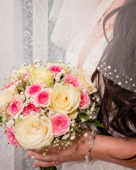 Close-up of a bride holding a floral wedding bouquet composed of cream and soft pink roses, complemented by small white filler flowers and green foliage, wrapped in a white ribbon. The bride's hand is