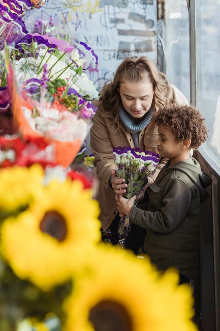 A woman and a young boy are inside a flower shop near a large window, engaging in a floral arrangement activity. The woman, with light brown, curly hair, is smiling warmly while she helps the boy, who